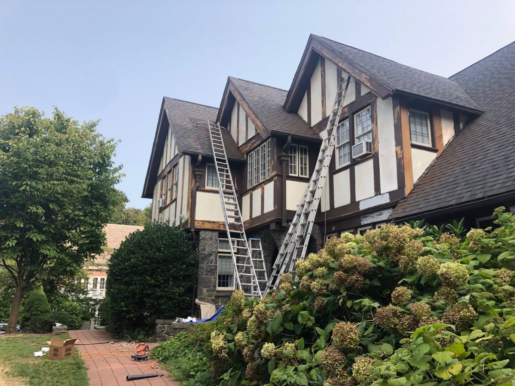 three eave home that has brown trim and tan siding. The brown trim is partially stripped and ready for repairs. there are ladders leaning against the house 